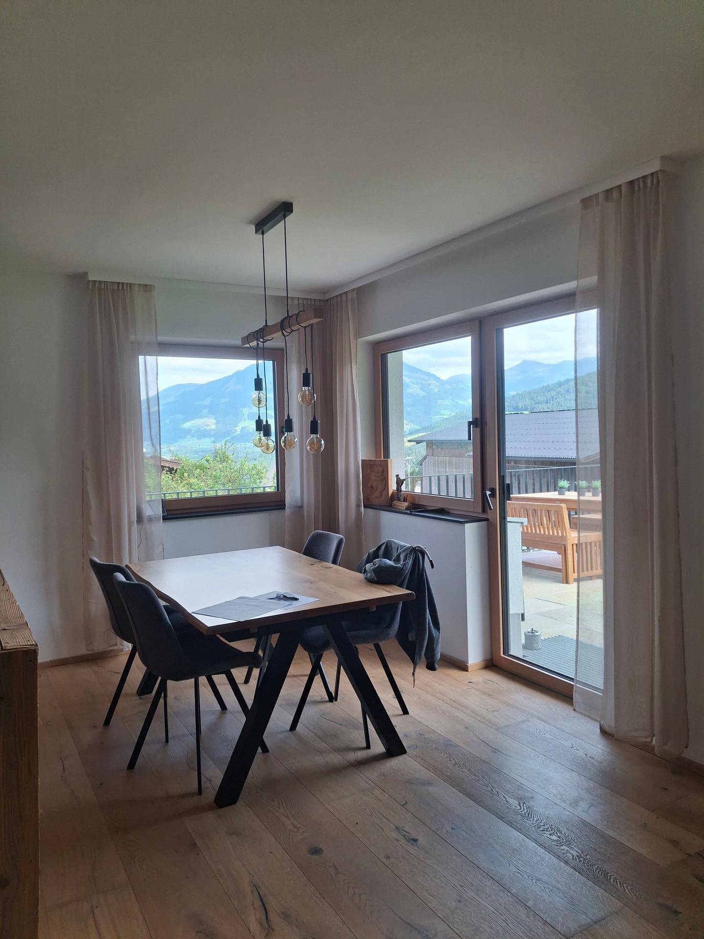 Essbereich mit Holztisch und Stühlen, großen Fenstern und Zugang zur Terrasse mit Bergblick.