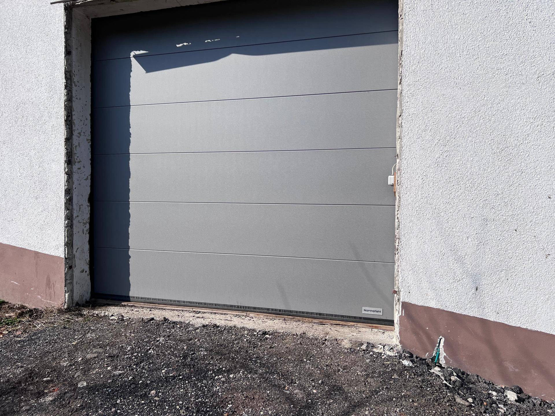 Nahaufnahme einer modernen, grauen Sektionaltür einer Garage mit weißer Fassade und Schotterboden.
