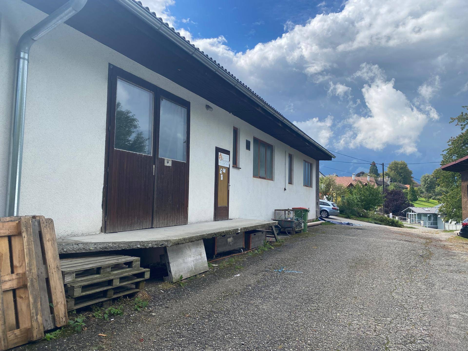 Seitenansicht des Gebäudes mit Laderampe, großen Holztüren und Schotterfläche davor unter bewölktem Himmel.