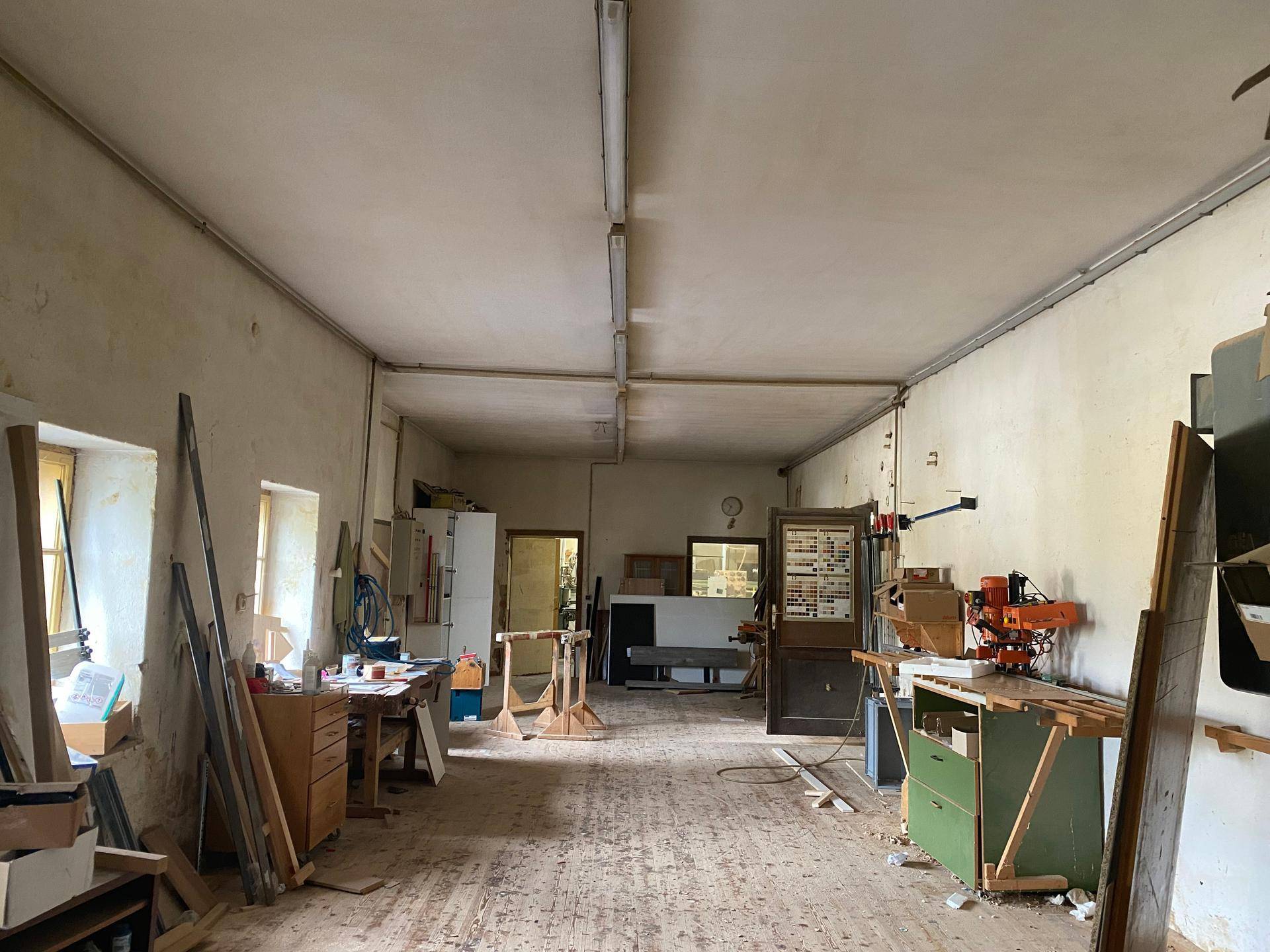 Längliche Holzwerkstatt mit Werkbänken, Maschinen und Fenstern, die den Raum mit Tageslicht füllen.