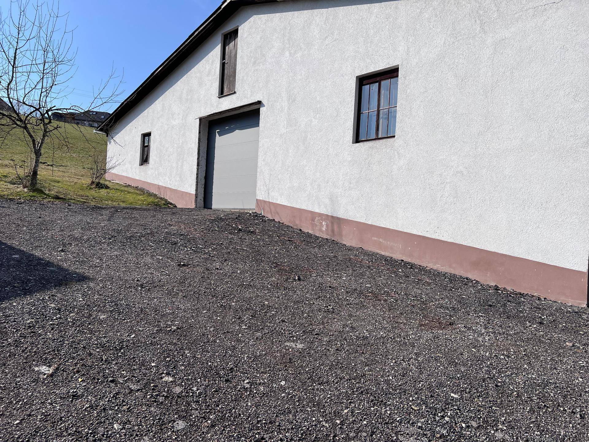 Rückseite des Gebäudes mit weißer Fassade, grauer Garagentür und Schotterauffahrt an einem sonnigen Tag.