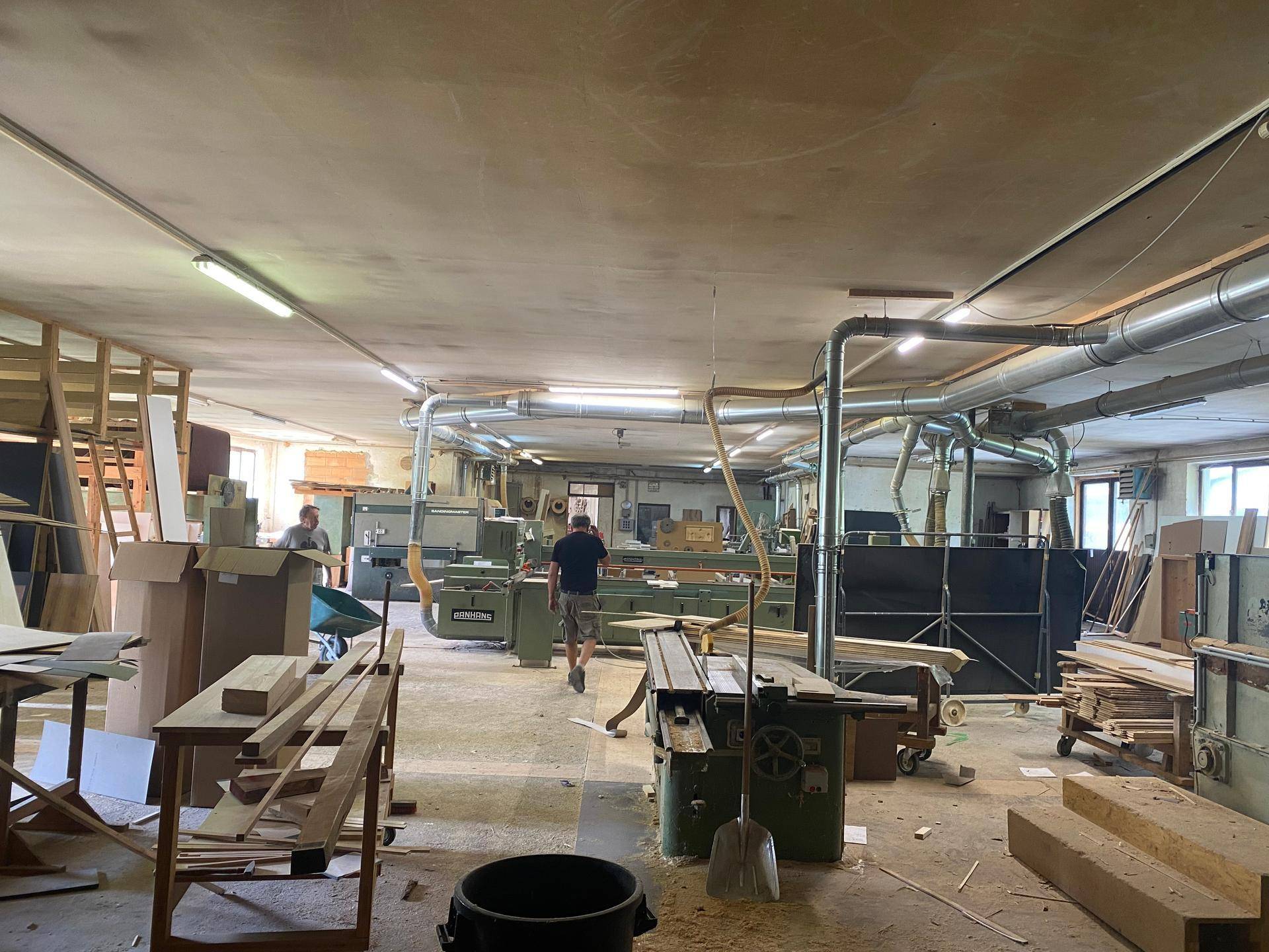 Weitläufige Holzwerkstatt mit verschiedenen Holzbearbeitungsmaschinen, Absauganlagen und Holzstapeln.
