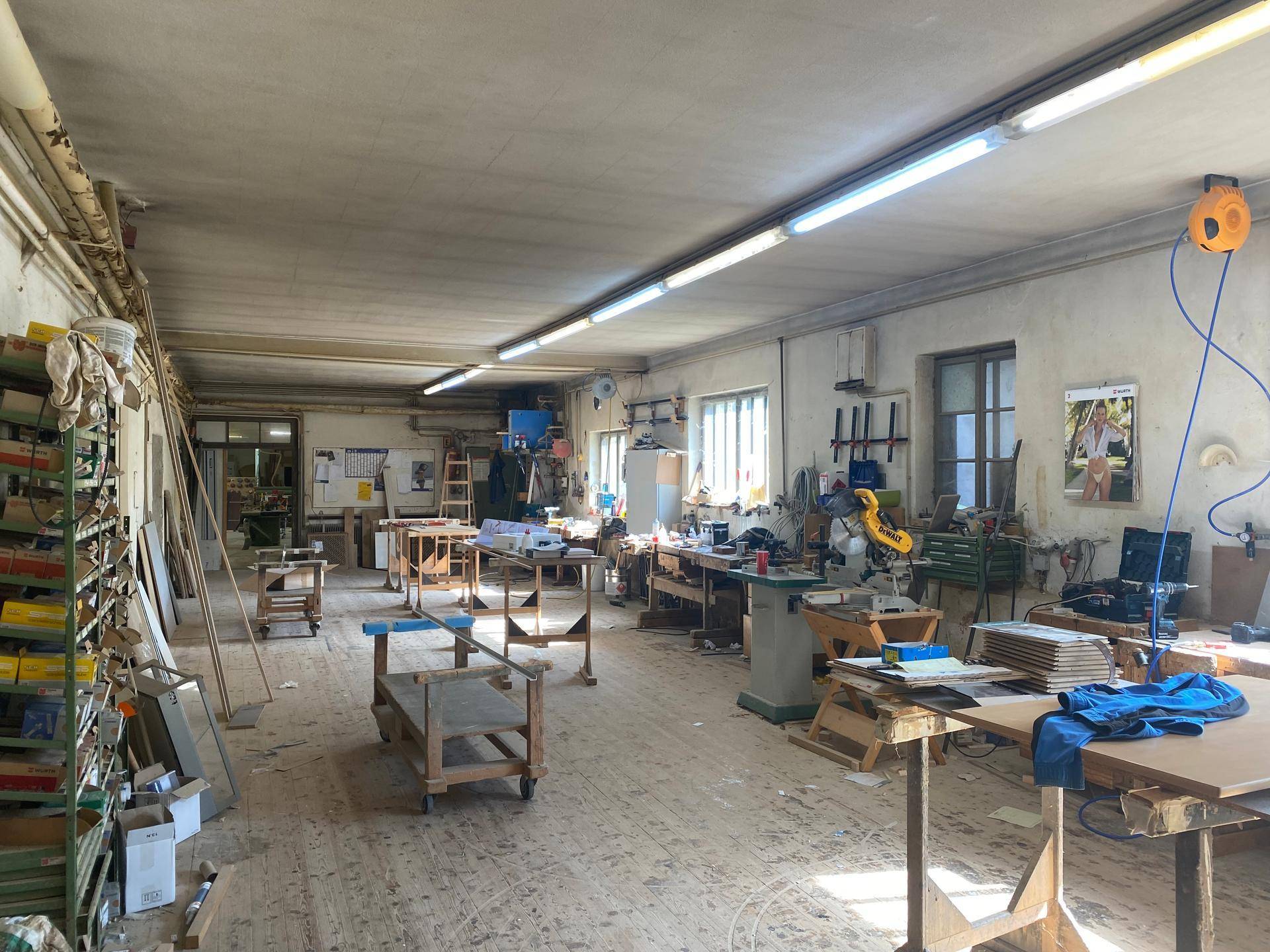 Helle Holzwerkstatt mit vielen Werkbänken, Maschinen und Fenstern, die Tageslicht hereinlassen.