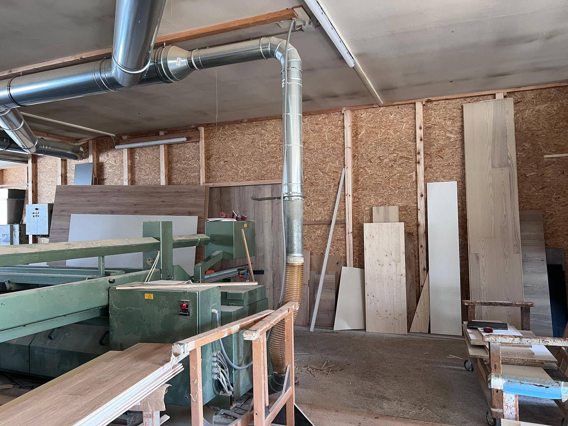 Detailansicht einer Holzbearbeitungsmaschine mit Absauganlage und gestapelten Holzplatten an der Wand.