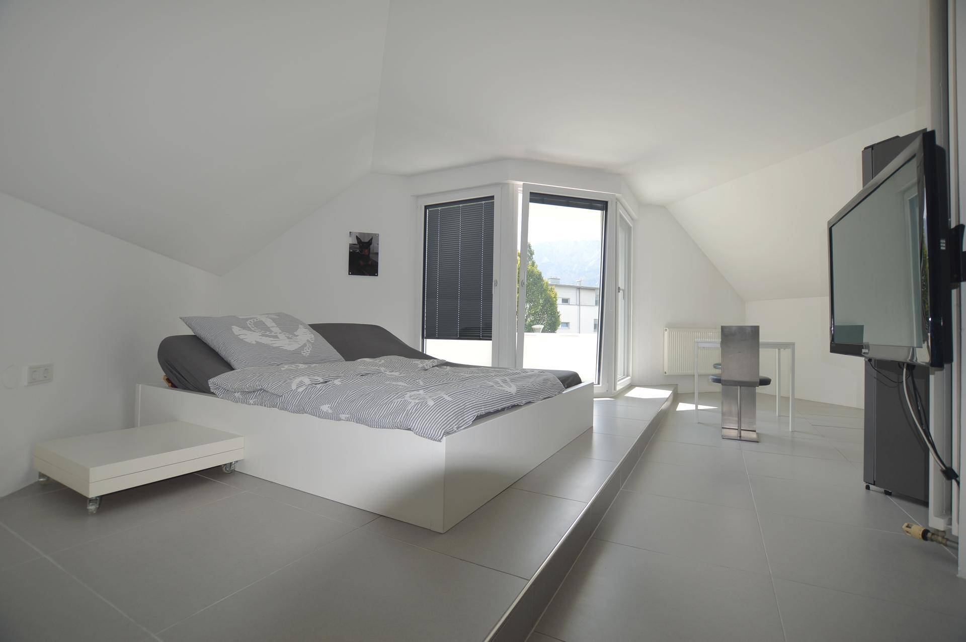 Modernes Schlafzimmer im Dachgeschoss mit erhöhtem Bettbereich und Zugang zum Balkon.