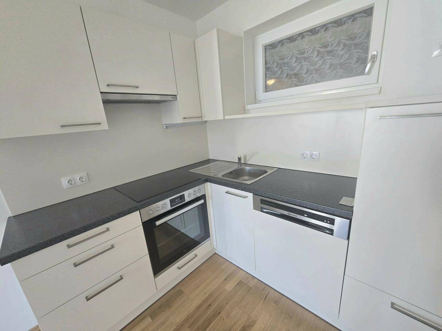 Funktionale Küche mit weißen Fronten, dunkler Arbeitsfläche und Holzfußboden, inklusive Backofen und Geschirrspüler.