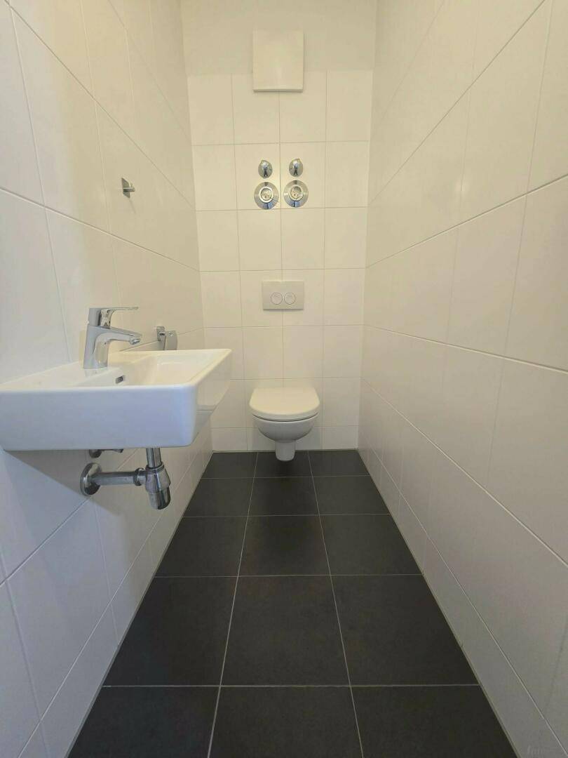 Gäste-WC mit weißen Wandfliesen, dunklem Fliesenboden und wandhängendem WC, inklusive Handwaschbecken.