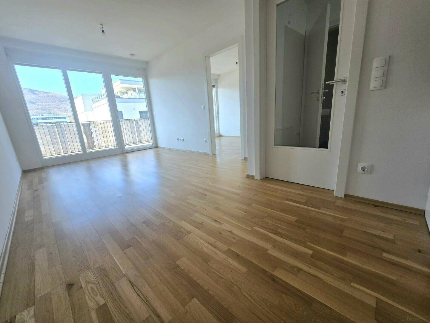 Heller Wohnraum mit großen Fenstern, Holzboden und Zugang zum Balkon, bietet einen weiten Blick auf die Berge.