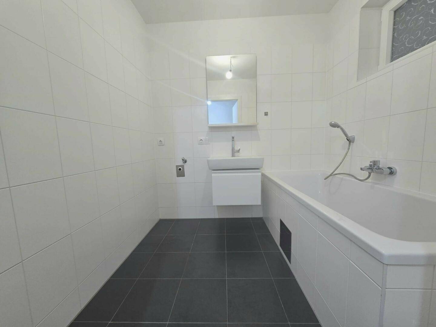 Modernes Badezimmer mit weißen Wandfliesen, dunklem Boden und Badewanne, ausgestattet mit einem Fenster.