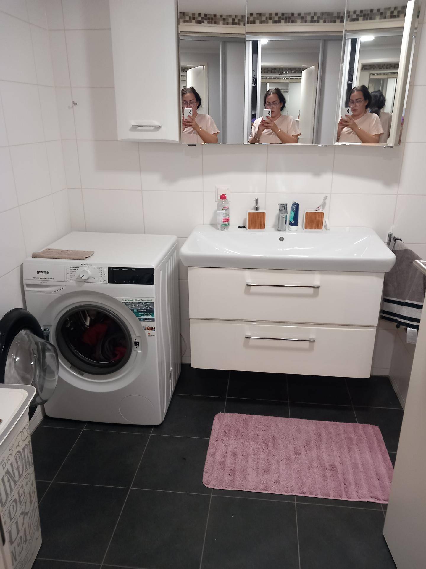 Badezimmer mit Waschmaschine, Waschbeckenunterschrank und Spiegelschrank über dunklen Fliesen.