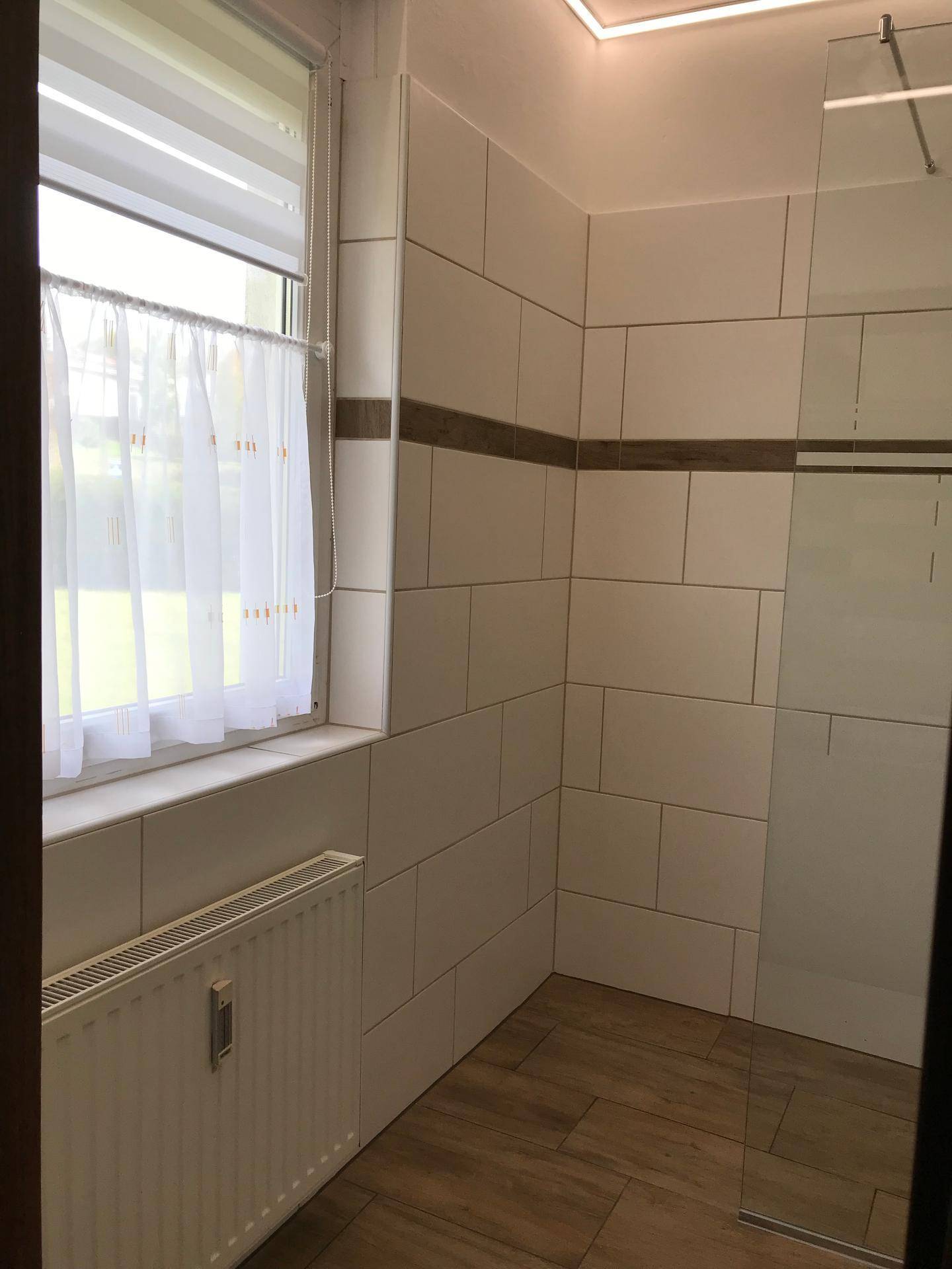 Helles Badezimmer mit weißen Fliesen, Holzoptikboden, Heizkörper und Fenster für natürliches Licht.