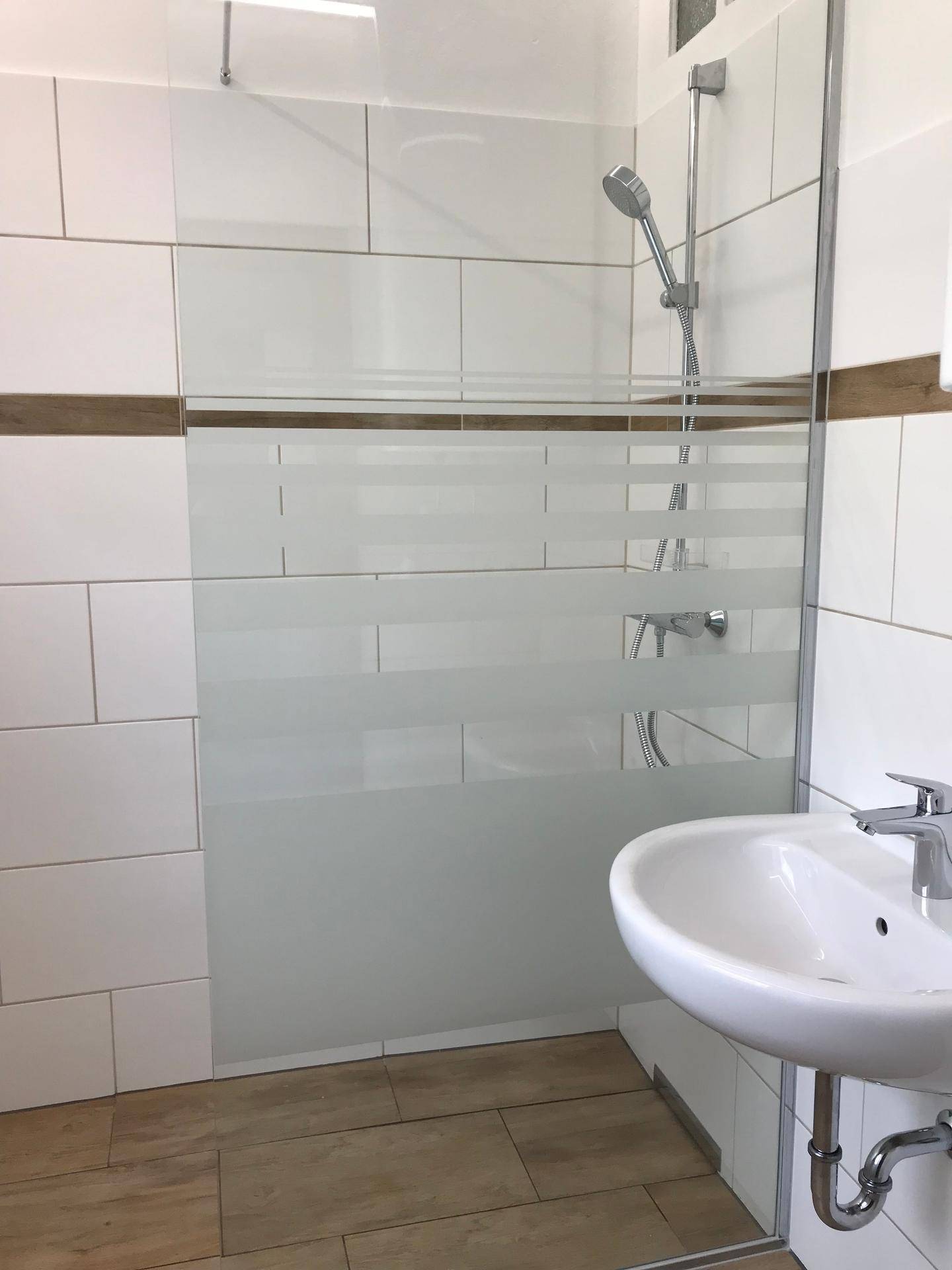 Modernes Badezimmer mit Duschkabine aus Glas und weißem Waschbecken auf Holzoptikboden.
