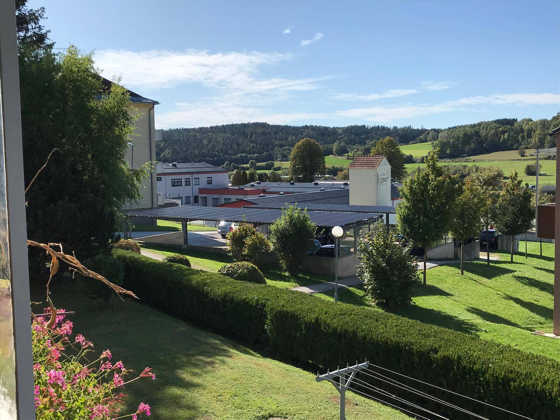 Weitläufige Grünflächen mit Bäumen, einem Carport und hügeliger Landschaft im Hintergrund unter blauem Himmel.