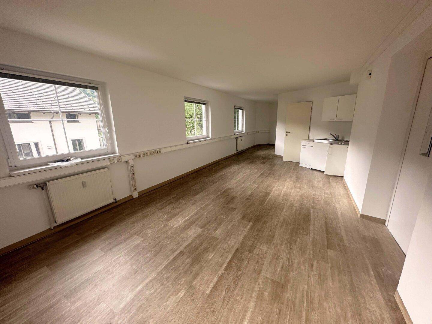 Heller Büroraum mit mehreren Fenstern, Laminatboden und einer kleinen Küchenzeile für Mitarbeiter.