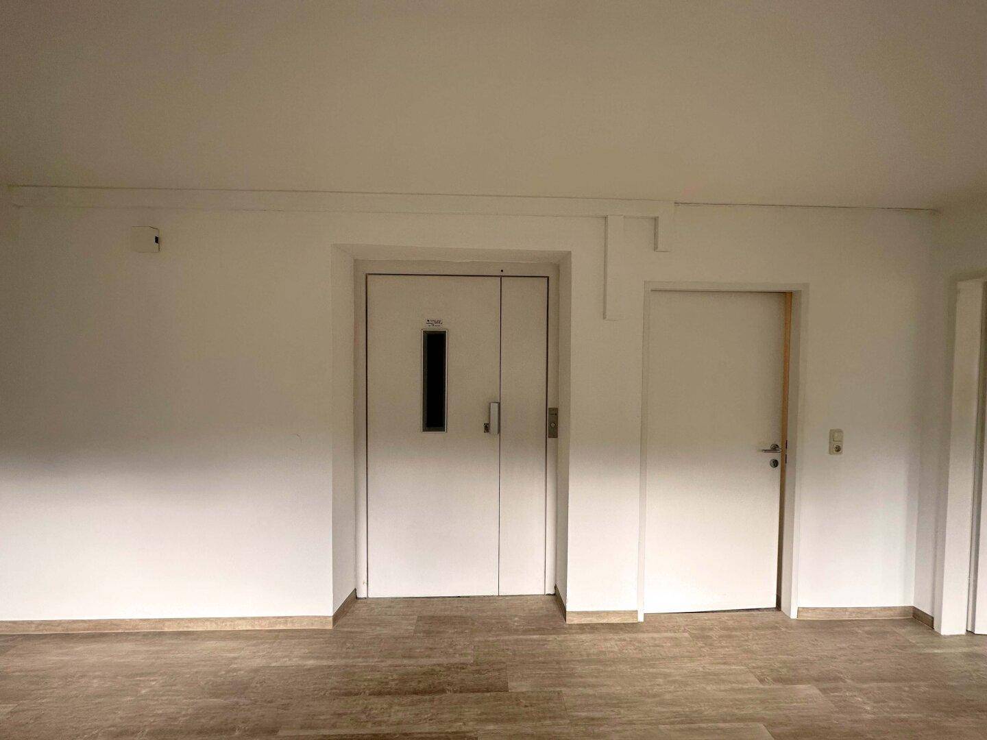 Heller Flur mit Laminatboden und zwei Türen, darunter ein Aufzug, der Zugang zu den Etagen bietet.