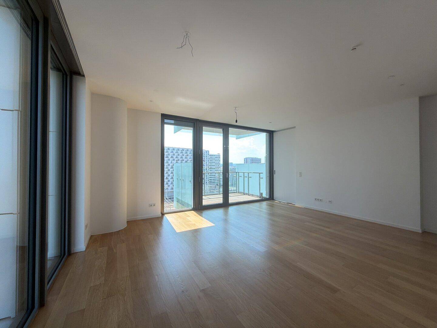 Geräumiges Zimmer mit viel Tageslicht, Holzboden und breiten Fenstern mit Blick nach draußen.