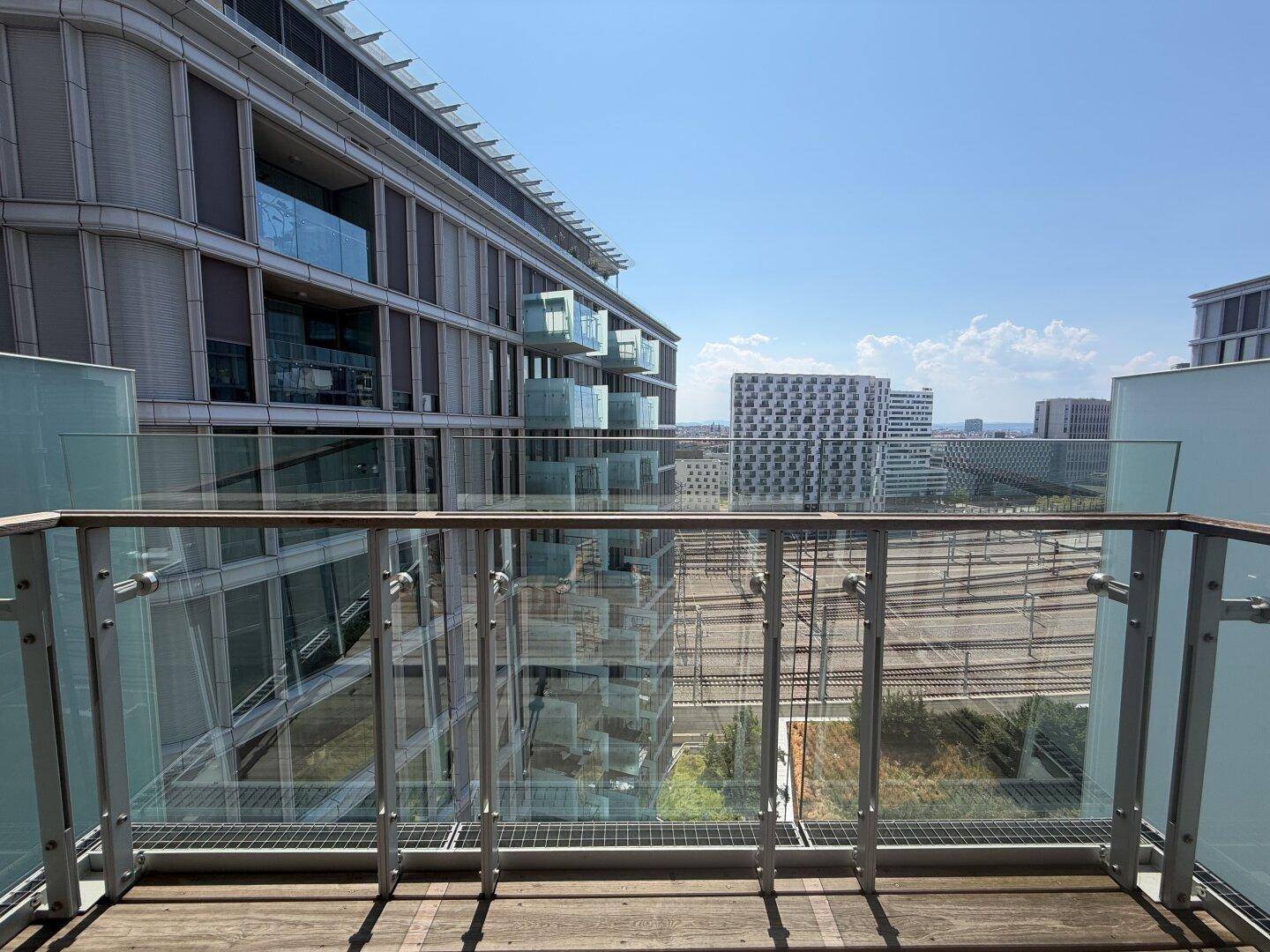 Balkon mit Glasgeländer und Holzboden, bietet einen Blick auf die Stadt und Bahngleise.