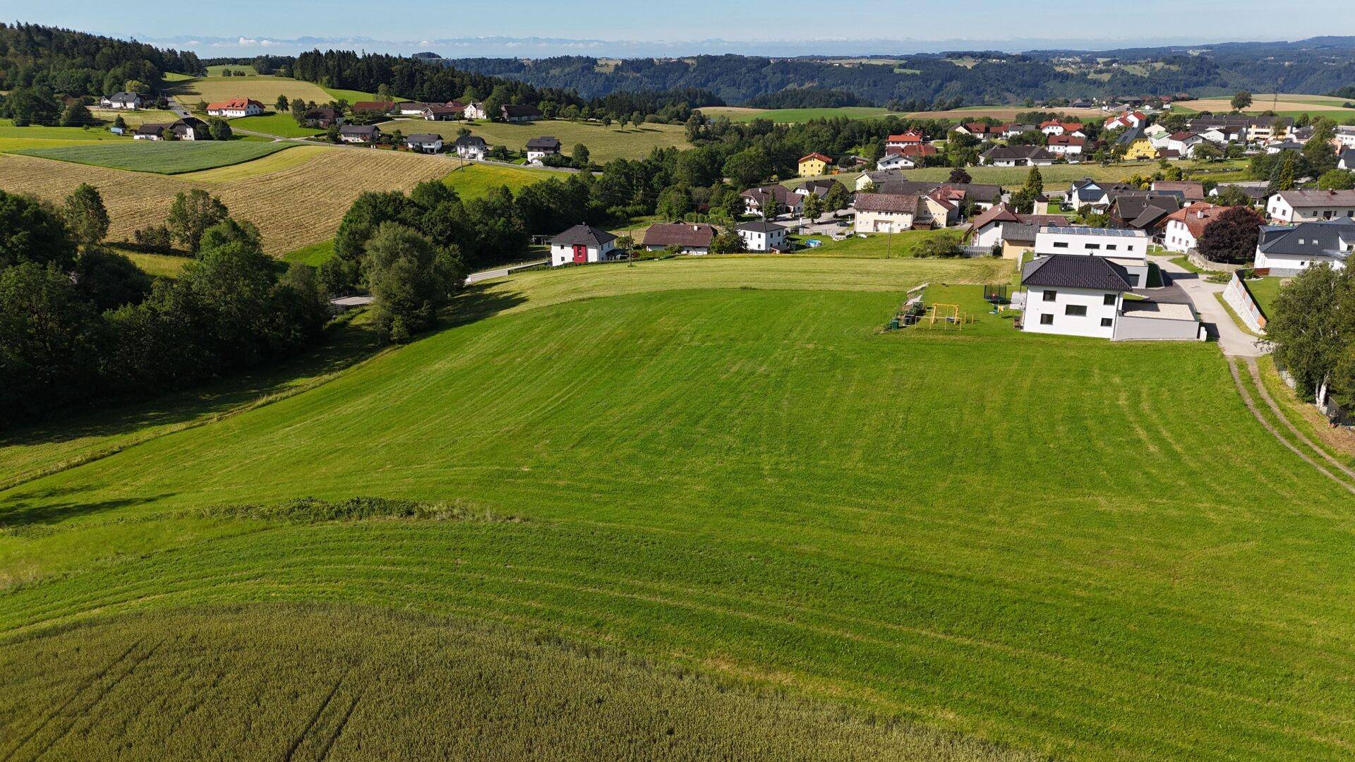 Panoramablick über sanfte Hügel, landwirtschaftliche Flächen und eine ländliche Siedlung unter blauem Himmel.