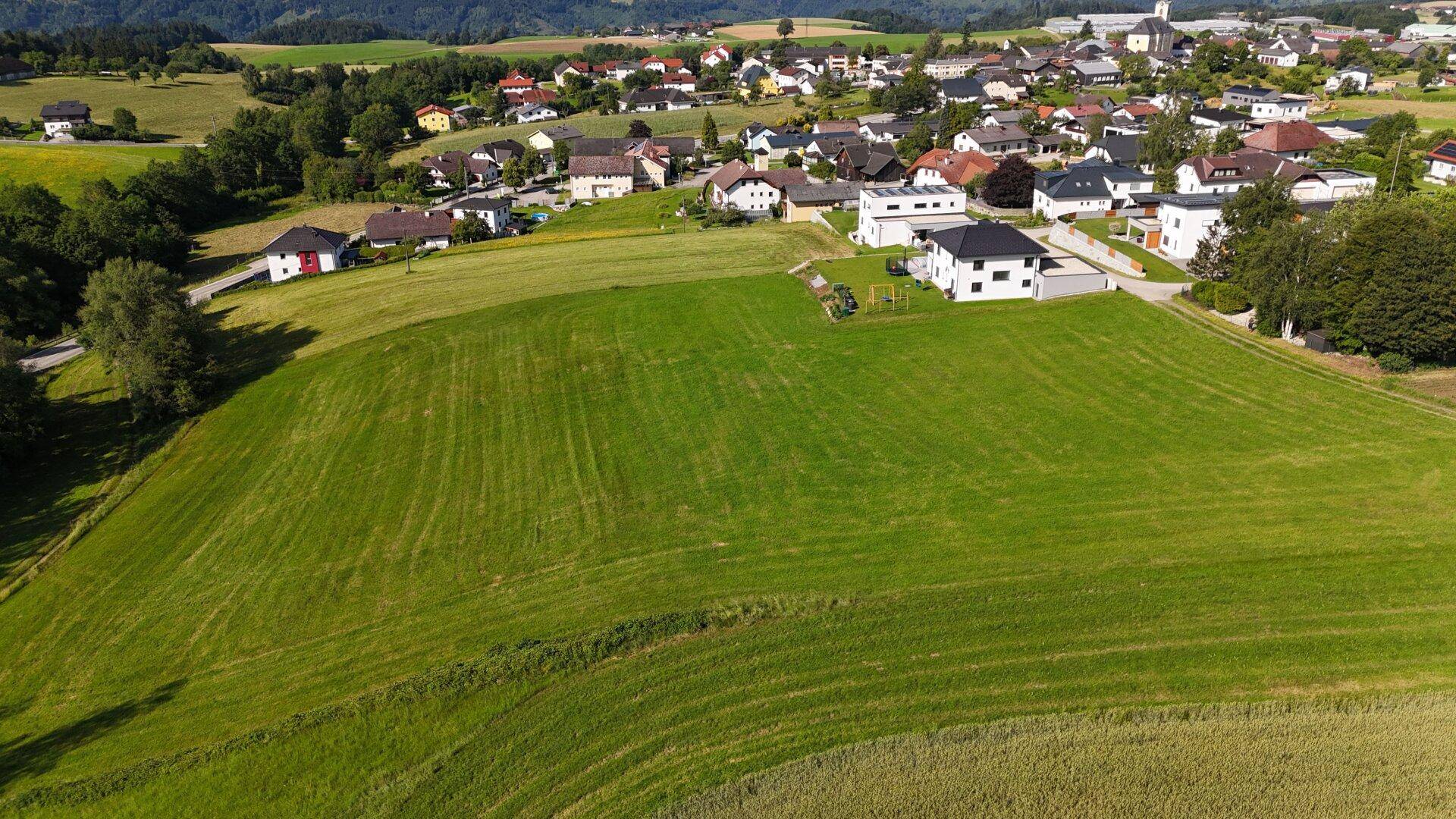 Blick von oben auf grüne Wiesen und Ackerland, umgeben von einer kleinen Ortschaft und Wäldern.