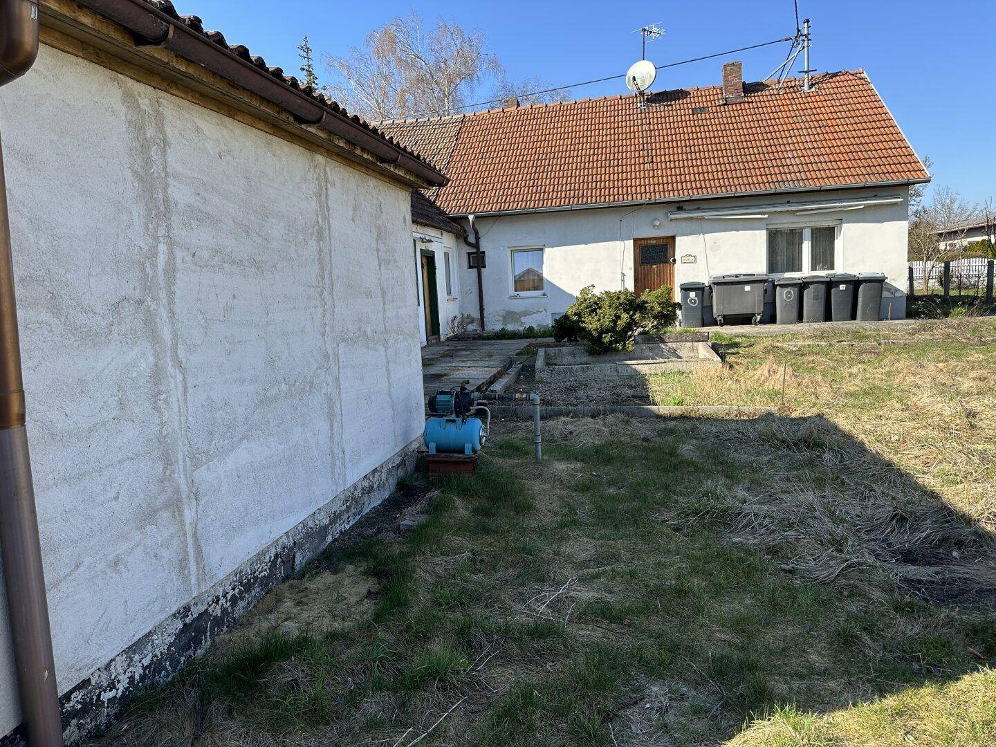 Rückansicht des Hauses mit gepflastertem Bereich, Mülltonnen und einer Wasserpumpe im Garten.