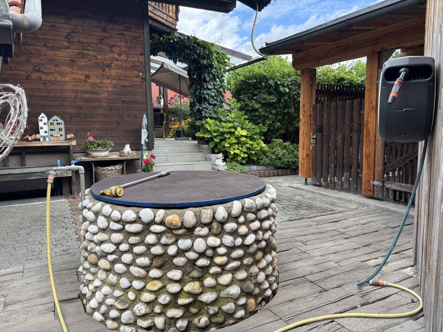 Rustikaler Gartenbereich mit Zierbrunnen, Holzterrasse und üppiger Bepflanzung.