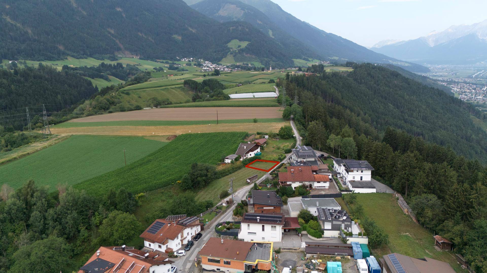 Luftaufnahme einer Dorfstraße mit Wohnhäusern, umgeben von grünen Feldern und Bergen.