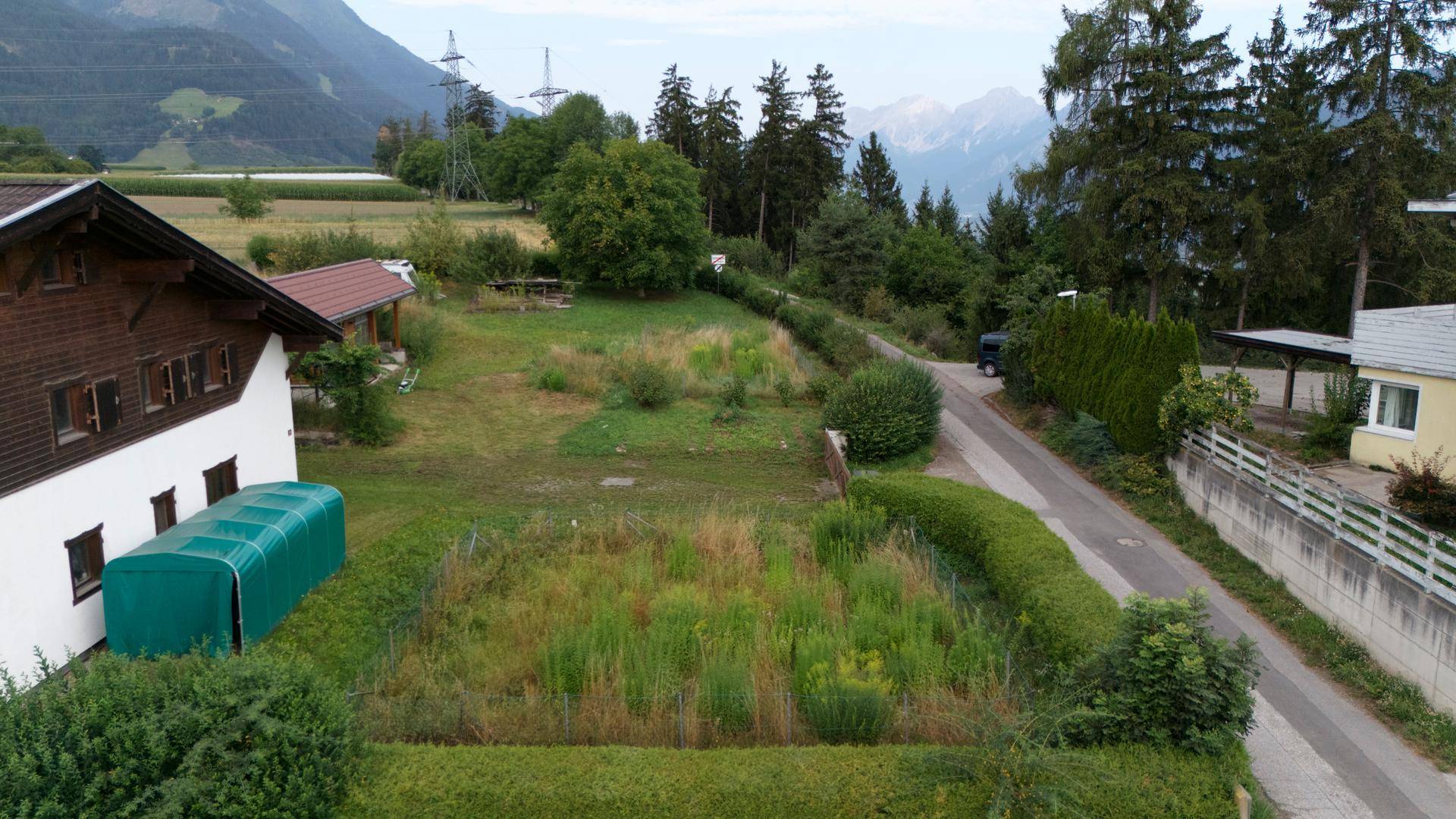 Ungepflegter Gartenbereich neben einem Haus mit Hecken und Blick auf die Berge.
