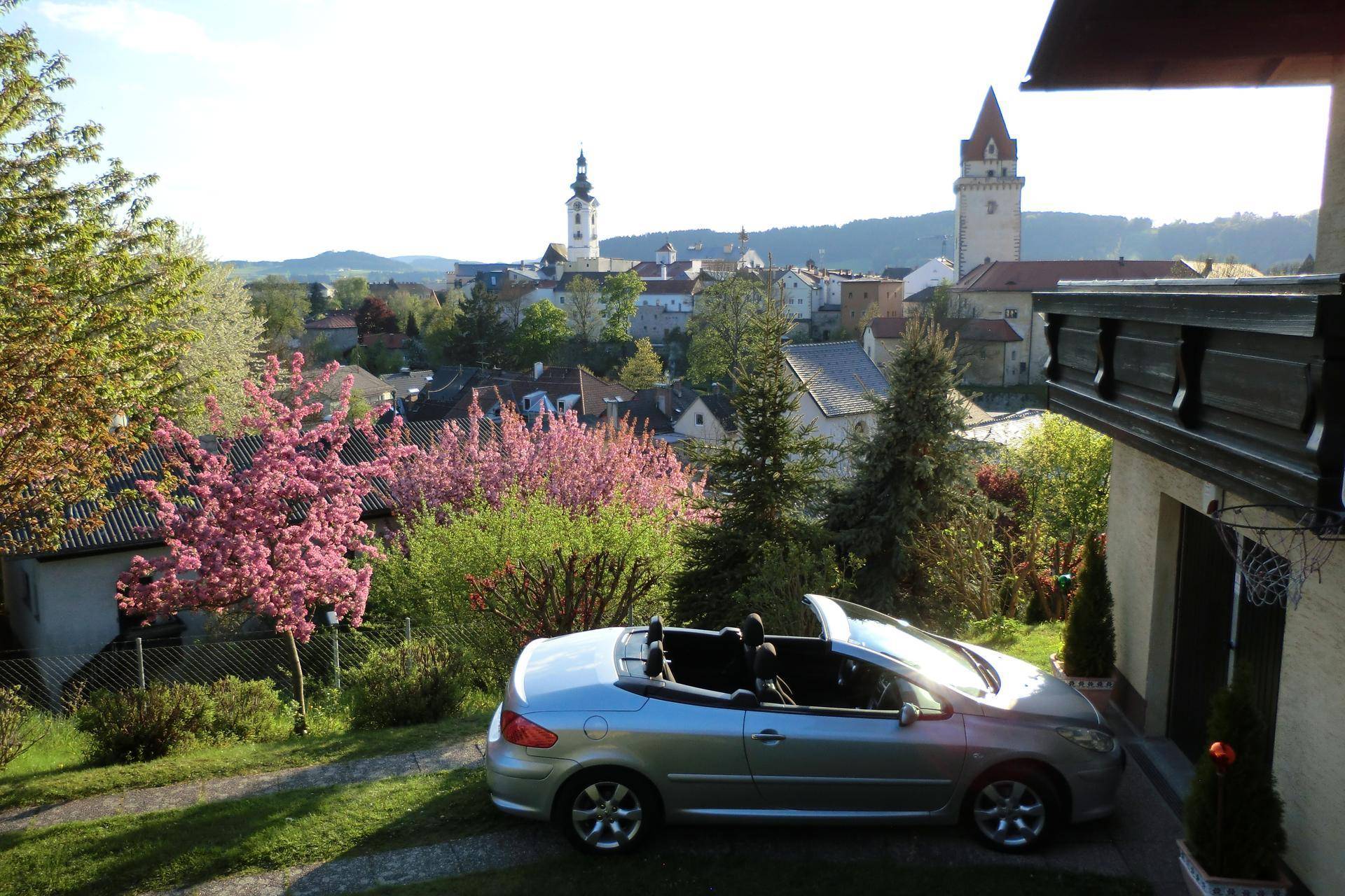 Blick vom Garten auf die Stadt mit blühenden Bäumen und einem geparkten Cabriolet.