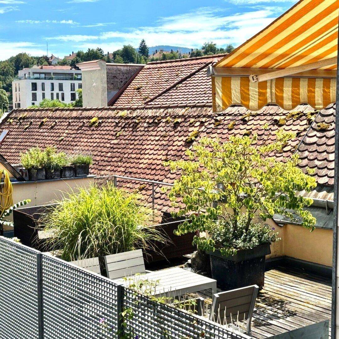 Sonnige Dachterrasse mit Pflanzen, Gartenmöbeln und Blick über die Dächer der Stadt.