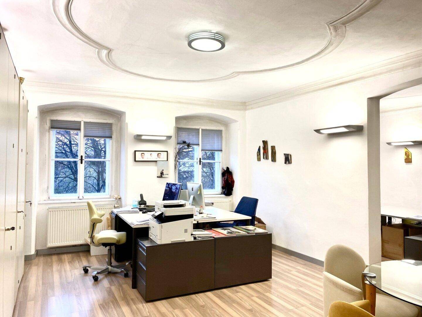 Geräumiges Büro mit Stuckdecke, großen Fenstern und mehreren Arbeitsplätzen.