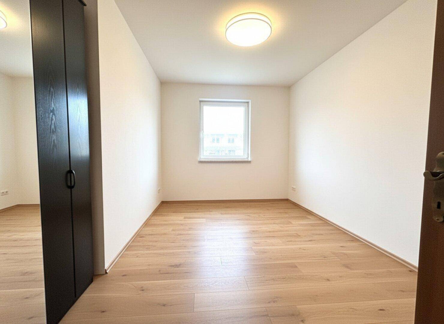 Helles, leeres Zimmer mit Holzboden und Fenster, ideal als Kinder- oder Gästezimmer.