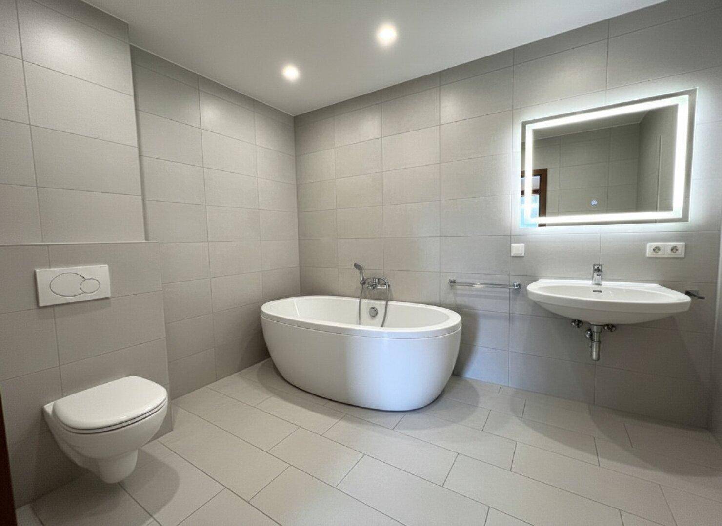 Modernes Badezimmer mit freistehender Badewanne, Wand-WC und beleuchtetem Spiegel über dem Waschbecken.