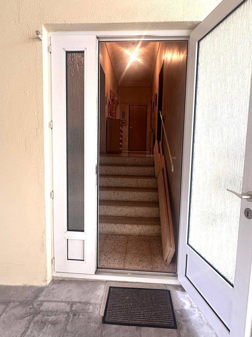 Eingangsbereich mit offener Tür, die zu einem Treppenhaus mit Terrazzoboden führt.