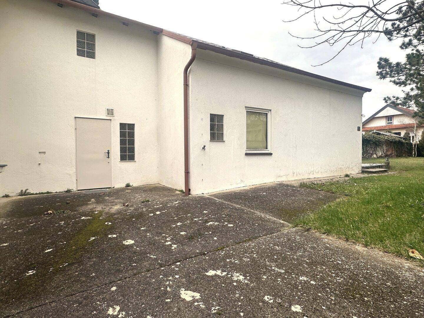 Seitenansicht eines Gebäudes mit weißer Fassade, kleiner Tür und gepflastertem Vorplatz.
