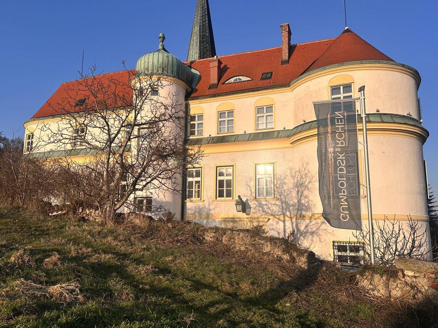 Historisches Gebäude mit Türmen, roten Dächern und einer Fahne auf einem grünen Hügel.