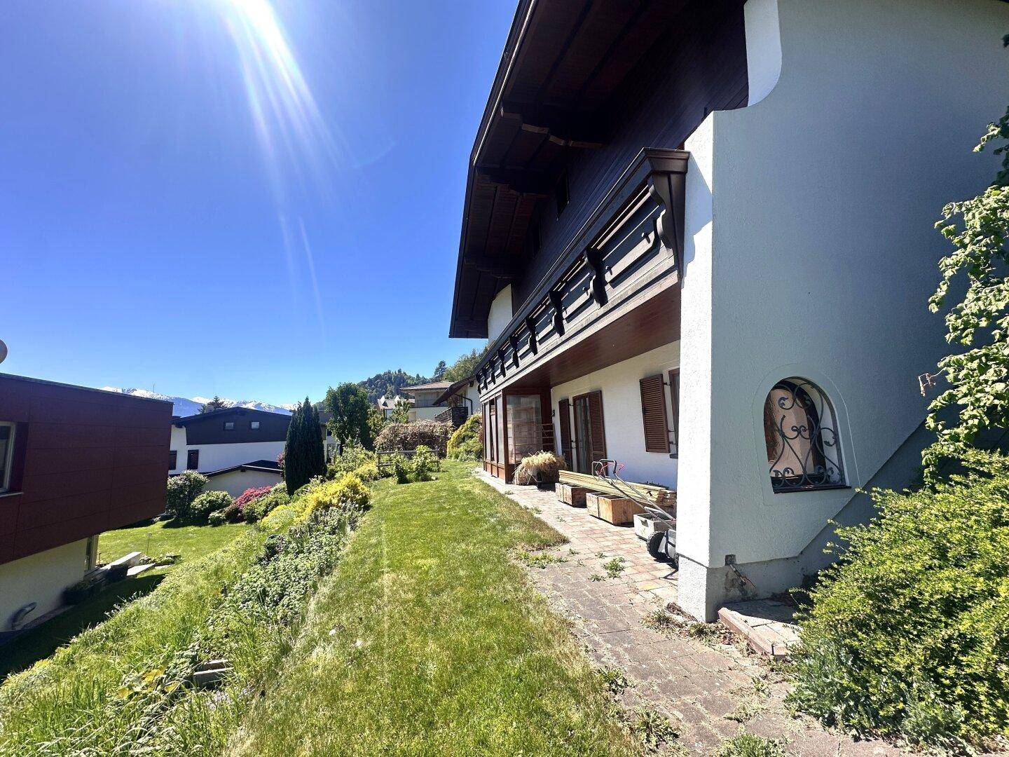 Seitenansicht des Hauses mit grünem Hang, kleiner Terrasse und Balkon unter strahlendem Sonnenschein.