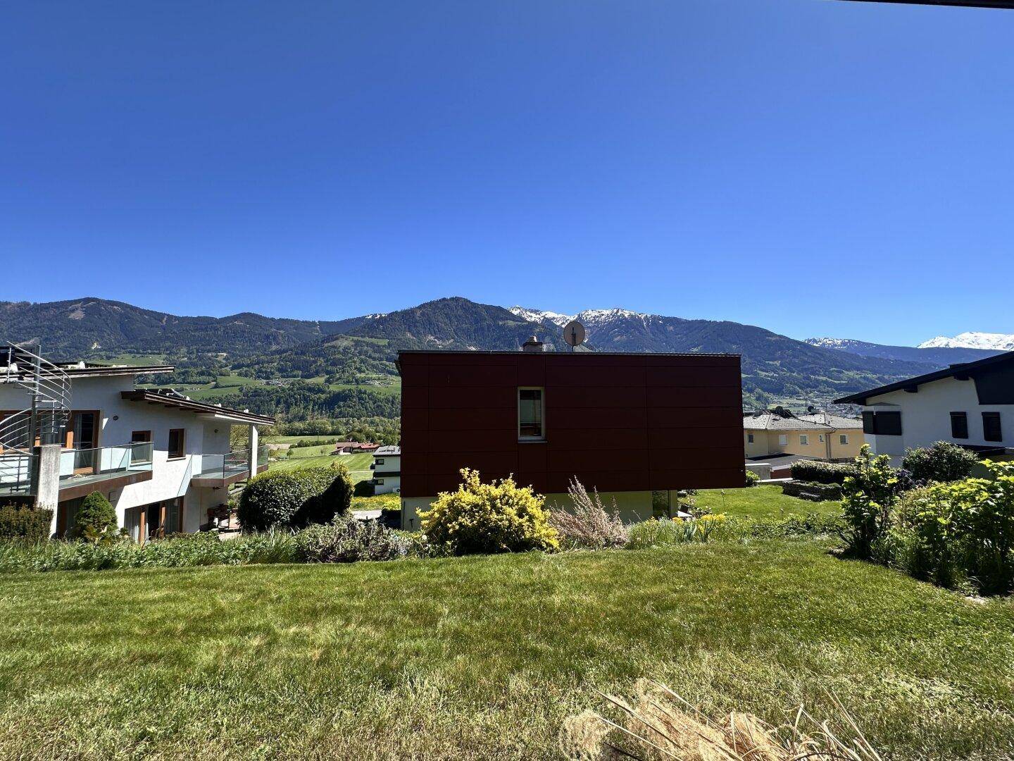 Breiter Ausblick auf grüne Wiesen, ein markantes rotes Gebäude und die ferne Bergkette unter blauem Himmel.