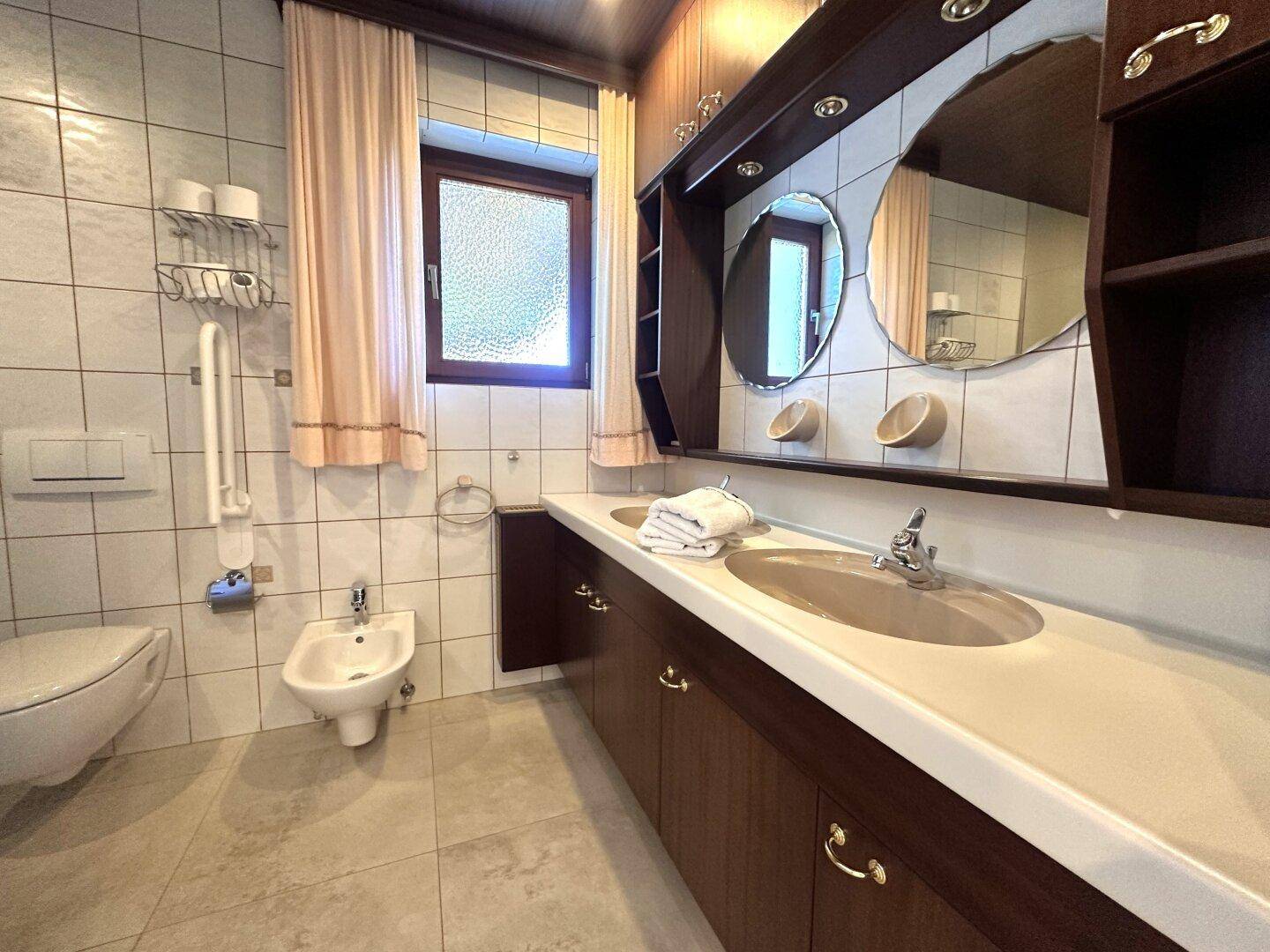 Funktionales Badezimmer mit Doppelwaschbecken, Toilette, Bidet und Fenster, ausgestattet mit Haltegriffen.