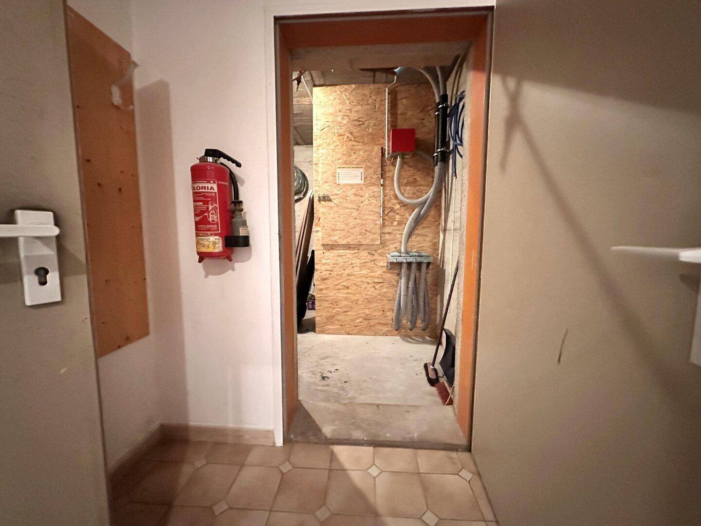 Lagerraum mit Fliesenboden und sichtbaren Installationen, ausgestattet mit einem Feuerlöscher.