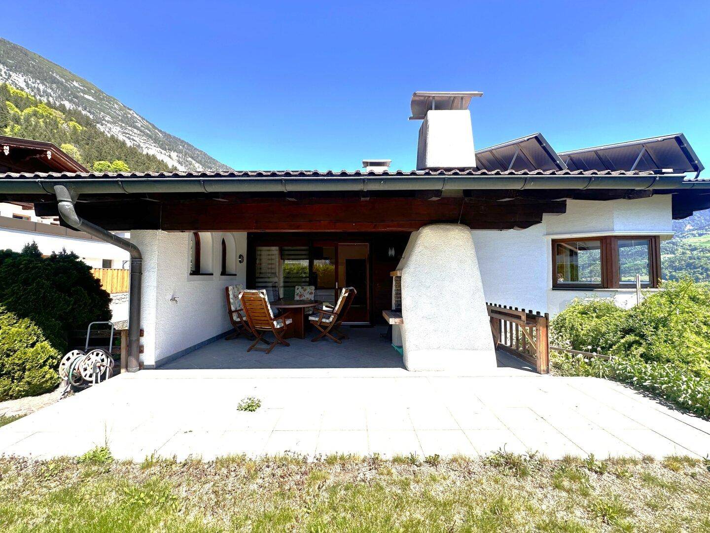 Rückseite des Hauses mit großer gepflasterter Terrasse, Rasenfläche und Panoramablick auf die Berge.