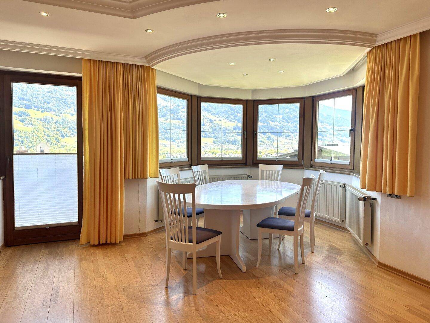 Heller Essbereich mit großem ovalem Tisch, umgeben von Erkerfenstern mit gelben Vorhängen und Bergblick.