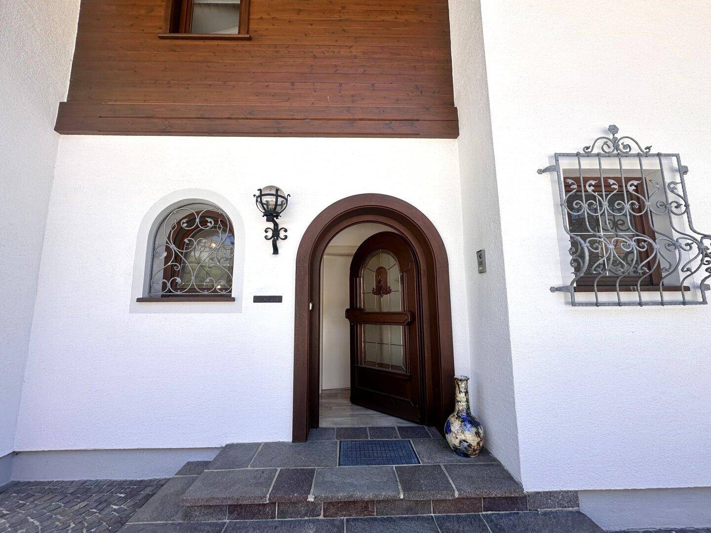 Eingangsbereich des Hauses mit rustikaler Holztür, Steinstufen und dekorativen Fenstergittern.