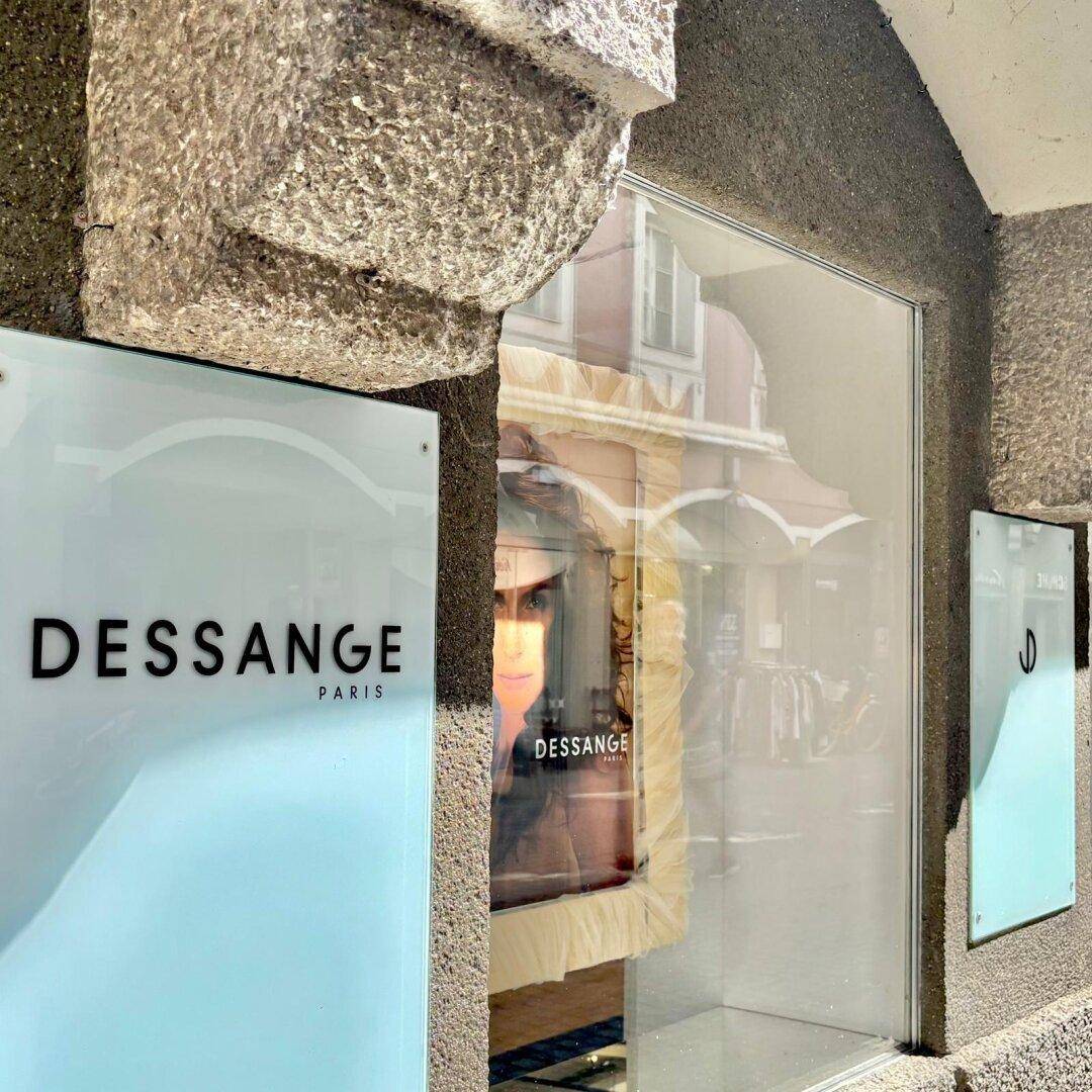 Detailansicht eines Schaufensters mit "DESSANGE"-Schild und Spiegelungen der gegenüberliegenden Gebäude in der Stadt.