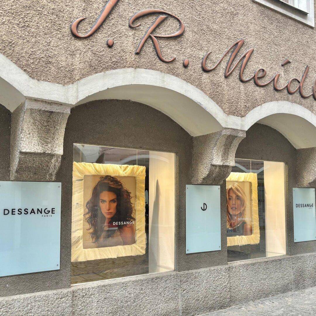 Nahaufnahme eines Schaufensters mit "DESSANGE"-Beschriftung und Werbeplakaten unter einem historischen Bogen.