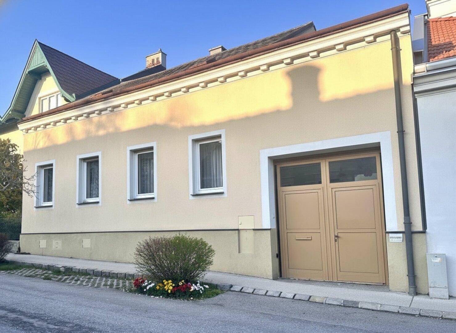 Traditionelles Haus mit beiger Fassade, weißen Fenstern und einem Garagentor zur Straße hin.