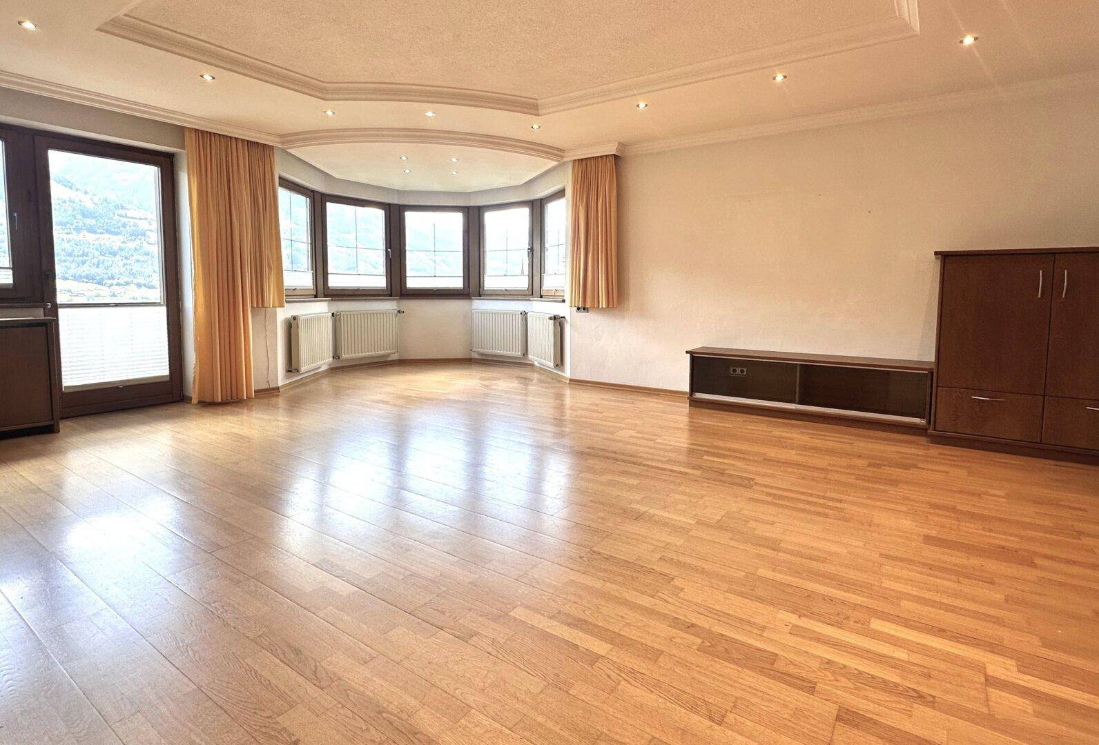 Geräumiges Wohnzimmer mit hellem Parkettboden, Erkerfenstern und einer dekorativen Decke.