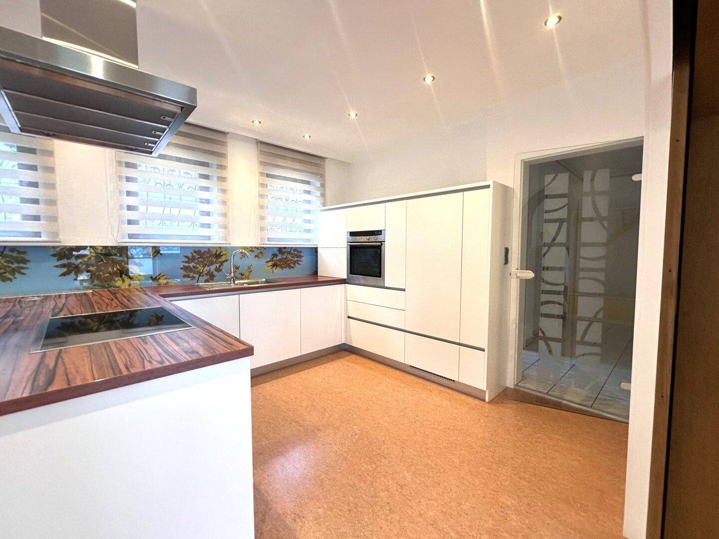 Moderne Küche mit weißen Schränken, Holzarbeitsplatte, Korkboden und einer Glastür zur Sauna.