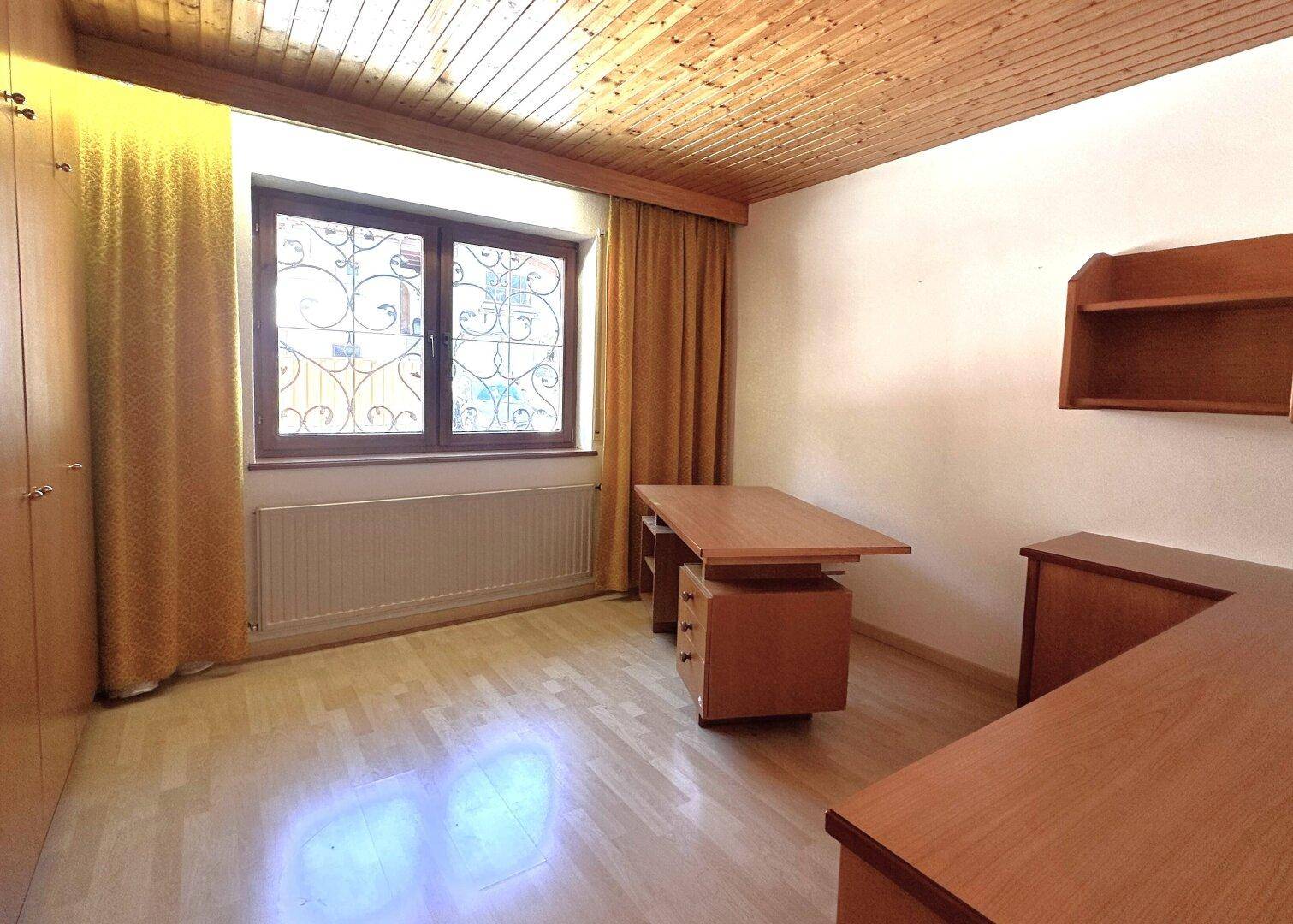 Gemütliches Arbeitszimmer mit Holzdecke, Schreibtisch und Fenster mit dekorativem Eisengitter.