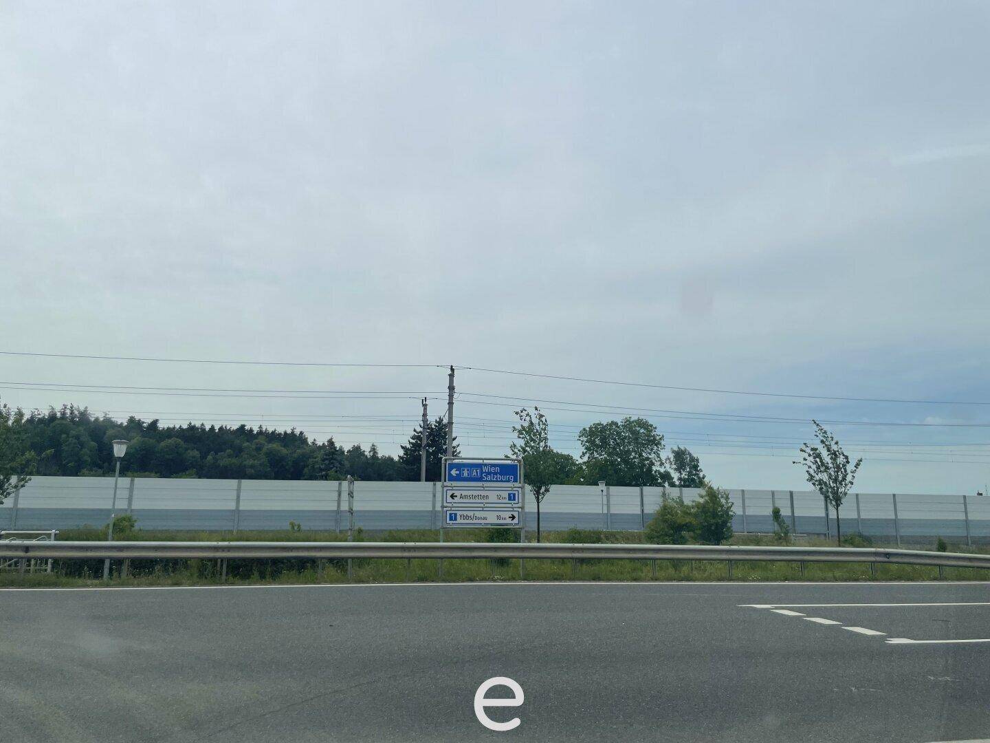 Blick auf eine Straße mit einer Lärmschutzwand und einem Verkehrsschild in der Ferne unter bewölktem Himmel.