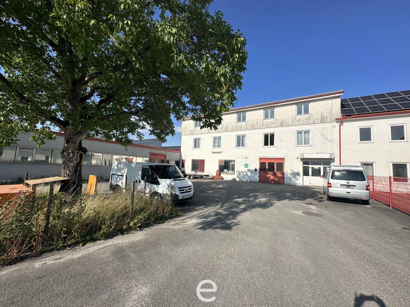 Rückansicht eines älteren Gebäudes mit Solarpaneelen auf dem Dach, umgeben von Bäumen und Parkplätzen.